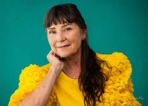 Dr. Lucas with dark hair and bangs wearing a yellow ruffled top, resting her chin on her hand and smiling.